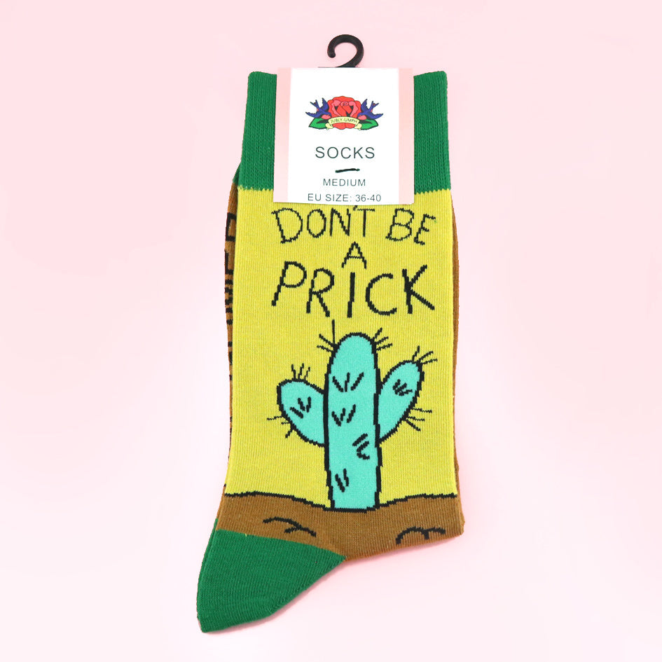 A pair of socks against a pink background. The socks are yellow, green and brown with a teal cactus. The socks read Don't Be A Prick.