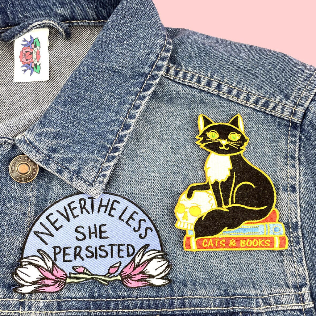 An iron on embroidered patch on a denim jacket. The patch is a black and white cat with a scull sitting on books. The patch reads Cat’s and Books. Another patch is also visible. It reads Nevertheless She Persisted.